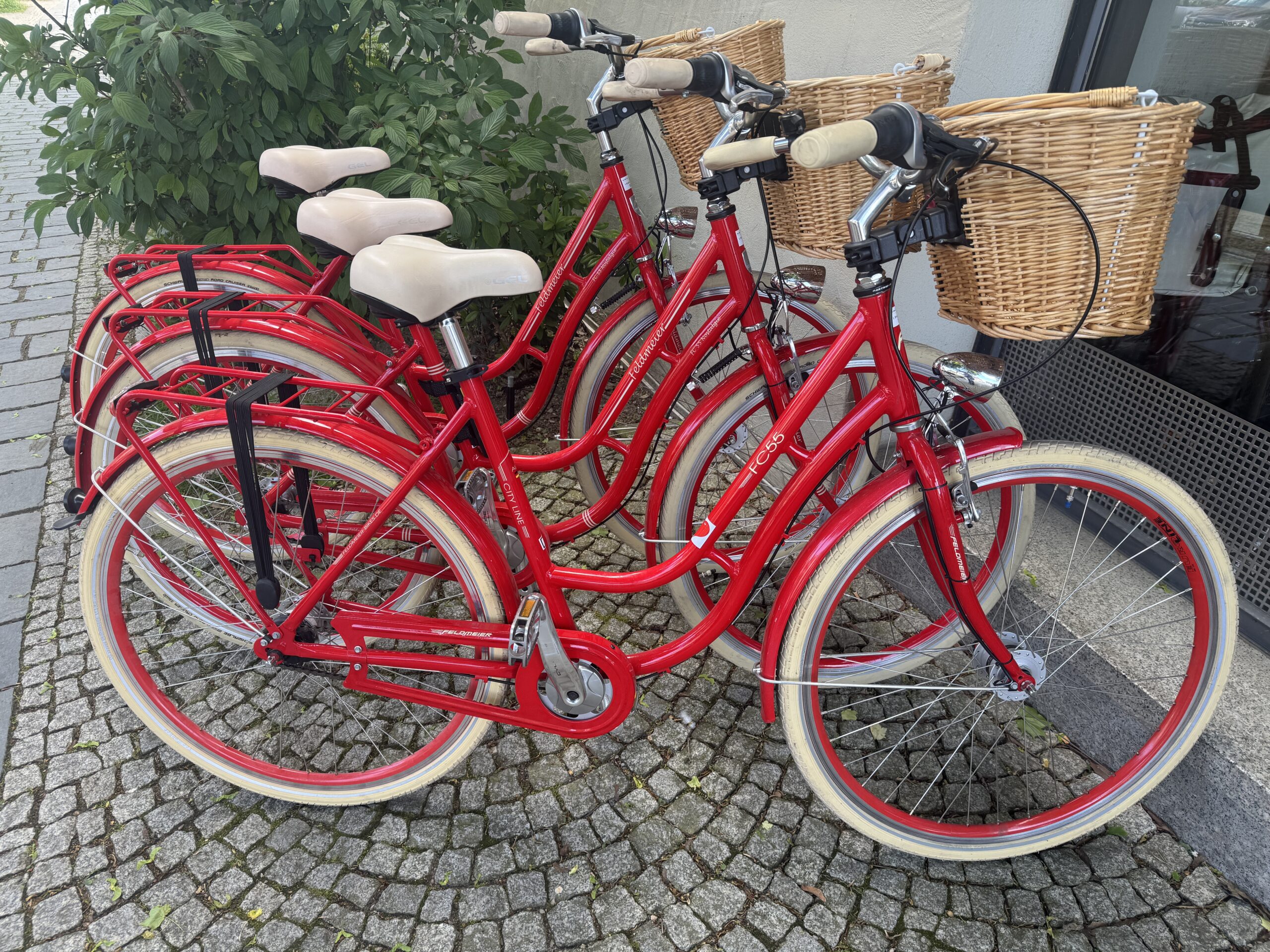 drei rote Stadtfahrräder mit tiefen Einstieg und Korb am Lenger vor grünen Busch auf Kopfsteinpflaster vor Hauswand